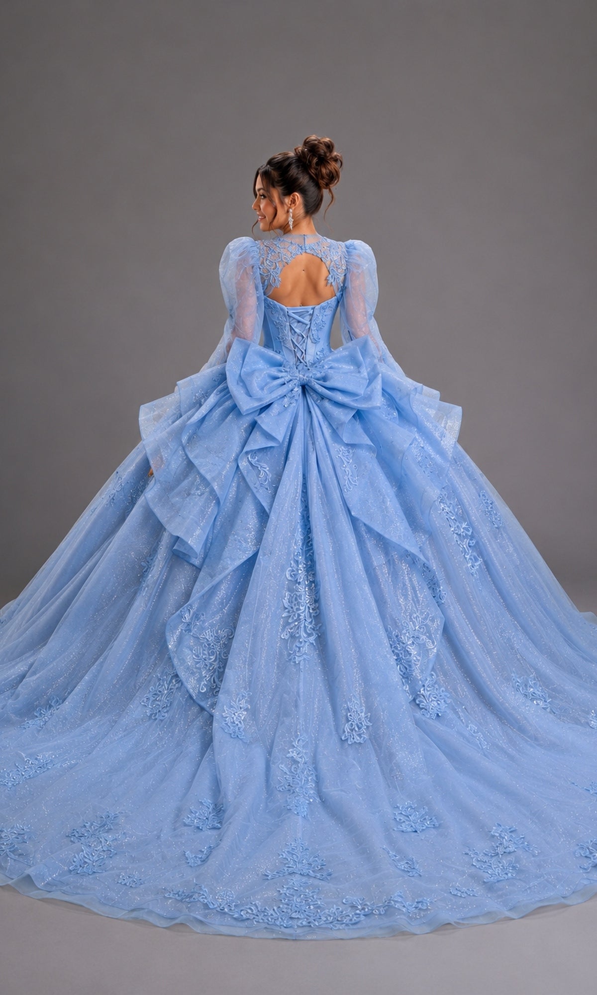 Back view of a light-blue Quinceanera Dress 5167 By Dancing Queen, featuring long sheer sleeves, a lace-up corset, and a voluminous, layered skirt with embroidered lace appliques and a large back bow.